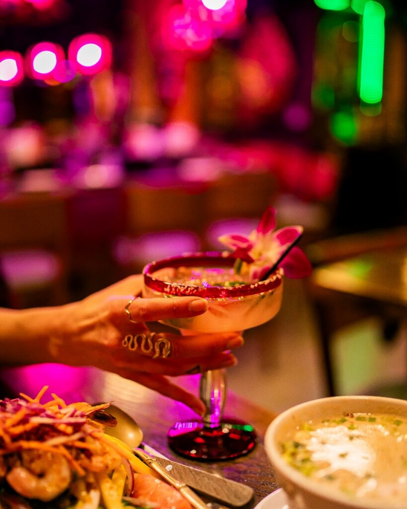 A hand with a gold snake ring holds a signature cocktail garnished with a pink orchid and a red-salted rim at Nicky’s Thai Kitchen, served alongside plates of authentic Thai food under vibrant pink restaurant lighting.