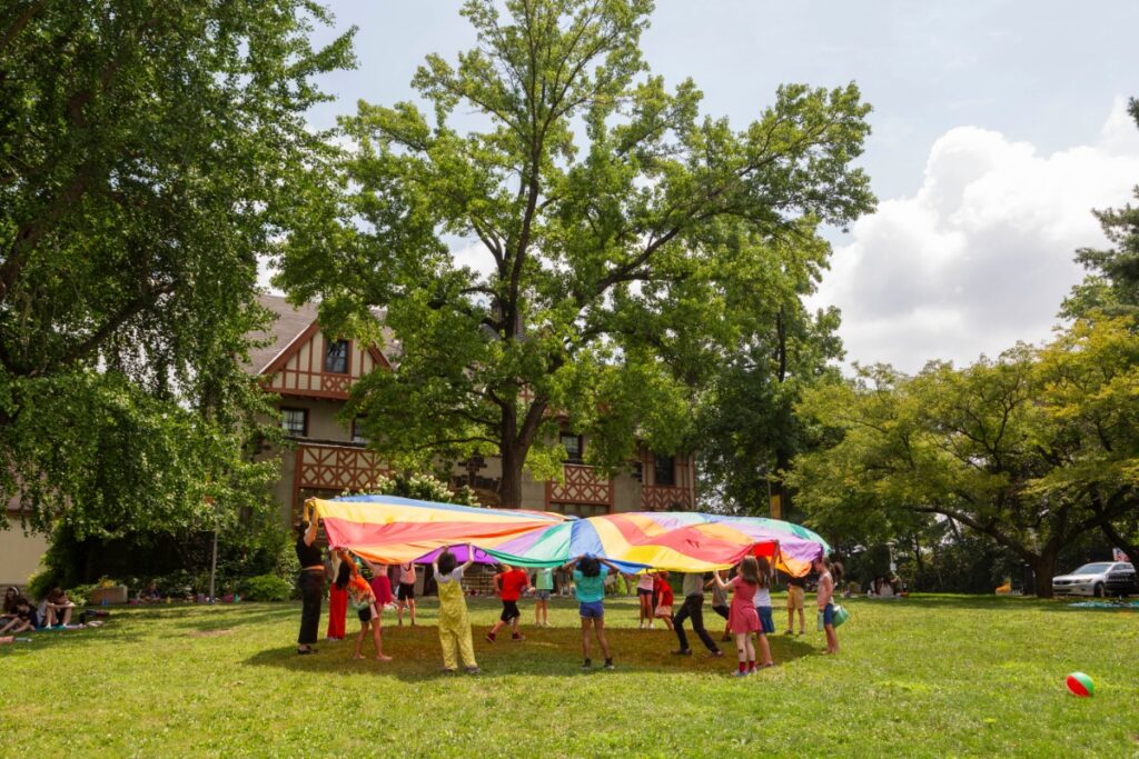 Children and instructors at PCA&M Summer Art Camp hold up a giant, colorful rainbow parachute on the grassy lawn of the Shadyside campus.