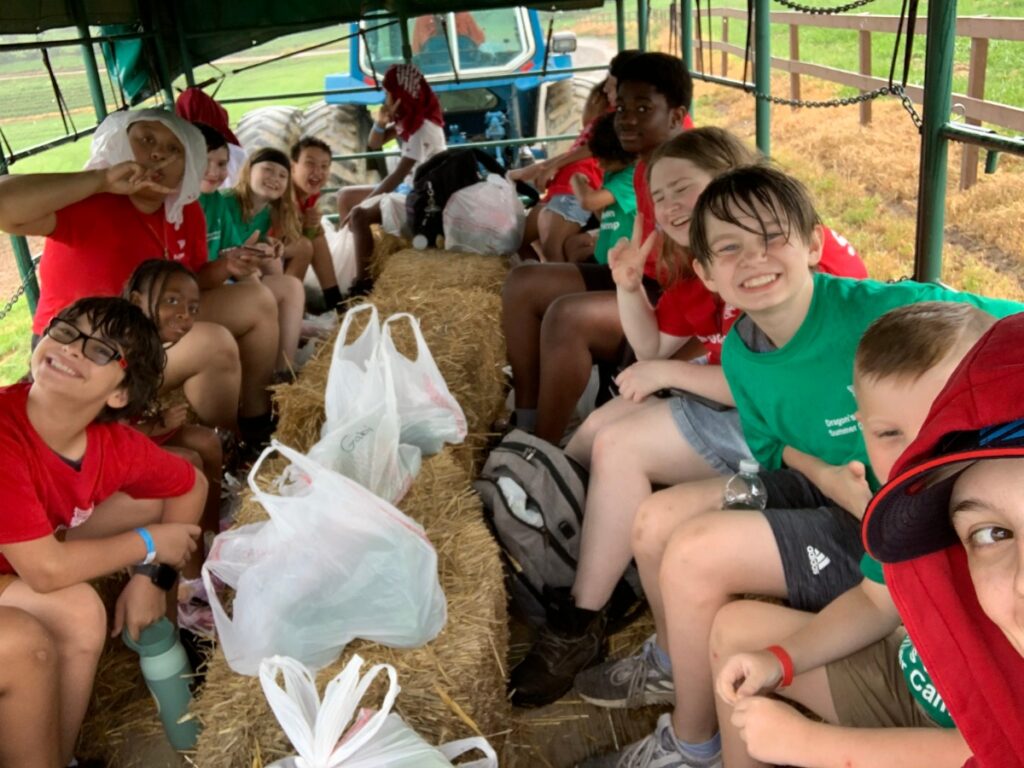 A group of smiling children in red and green camp t-shirts enjoy a hayride on a tractor-pulled wagon during a Dragon’s Den Summer Camp field trip in Pittsburgh.