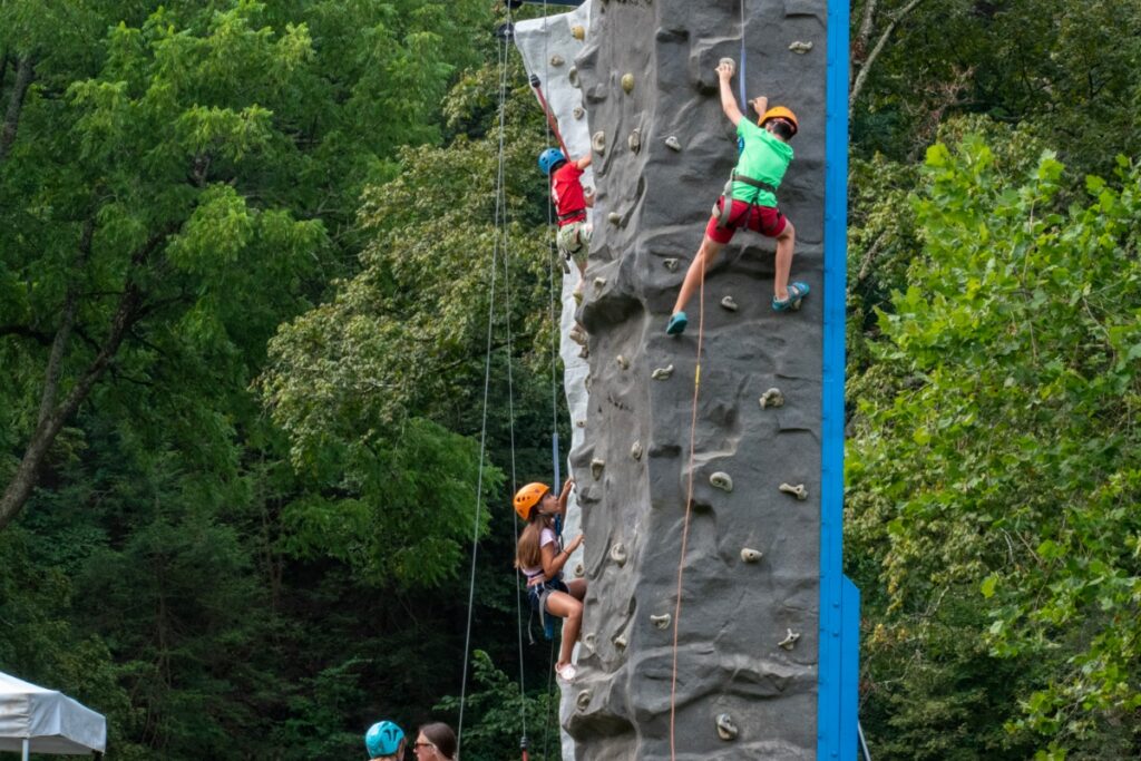 A young camper wearing a safety helmet and harness successfully scales a tall outdoor climbing wall at Camp Deer Creek, a traditional summer day camp in Pittsburgh.