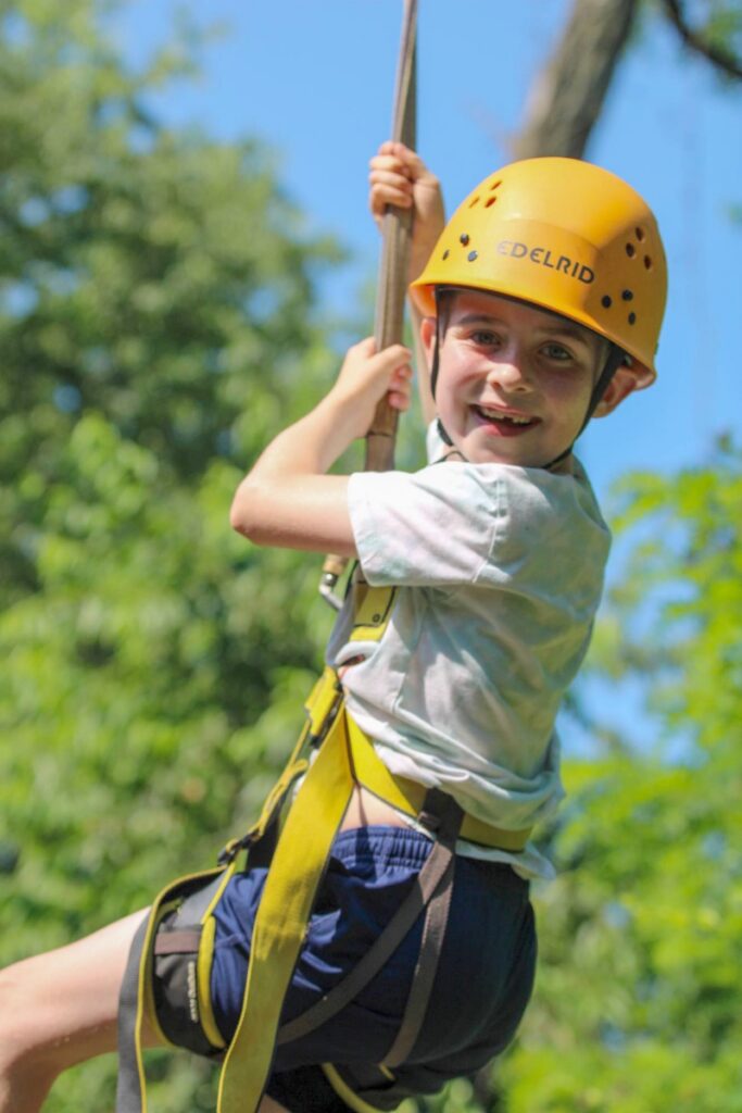 A smiling young boy wearing a yellow Edelrid safety helmet and climbing harness navigates a ropes course at JCC Day Camps, highlighting outdoor adventure and personal development for campers.