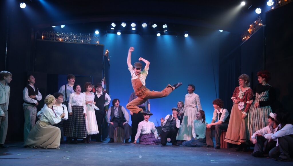 A young male performer in overalls performing a high leap on stage during a musical theater production at PMT Conservatory Summer Camp, surrounded by a cast in period-style costumes.