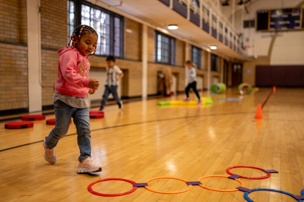 A young girl with beaded braids participates in a structured obstacle course on a gym floor during the Trinity Montessori & Reggio-Inspired Summer Program at Carlow University's Campus Lab School, while other children follow behind.