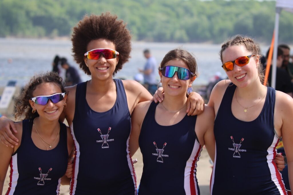 Four smiling young women in Three Rivers Rowing Association uniforms and athletic sunglasses stand together by the water, highlighting the teamwork and camaraderie of the youth rowing summer camp in Pittsburgh.