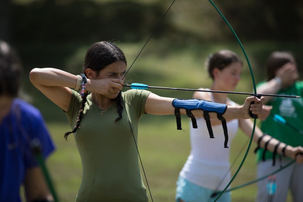 A young girl in a green t-shirt aims a bow and arrow with focused concentration during an archery lesson at Camp Twin Creeks, a co-ed sleepaway summer camp in the Allegheny Mountains.
