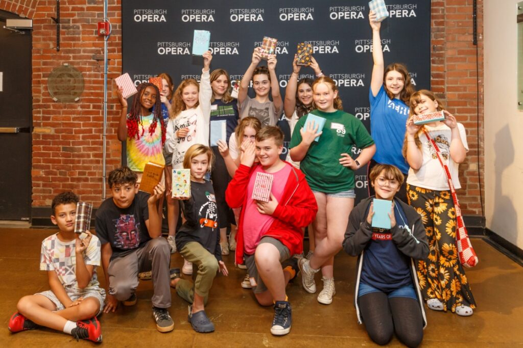 A diverse group of middle school students at Pittsburgh Opera's Bravo Academy proudly hold up their handmade costume or set design boxes in front of a Pittsburgh Opera backdrop at the Bitz Opera Factory.