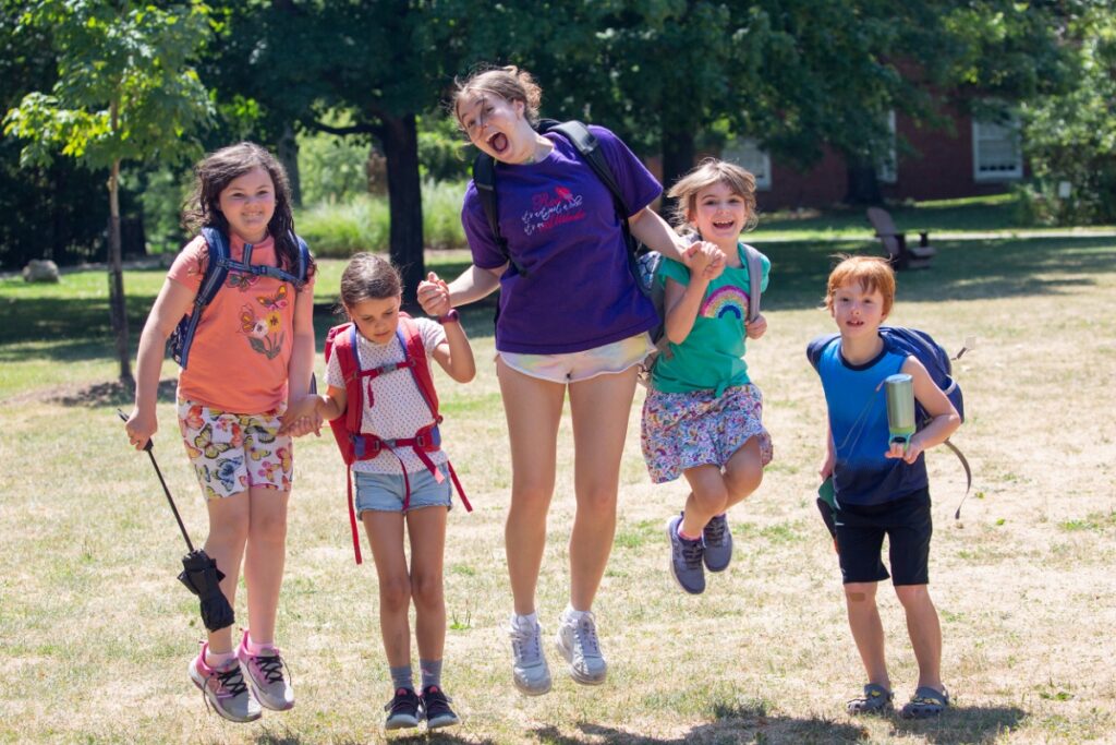 A group of diverse young campers and a counselor in a purple Chatham University t-shirt jumping joyfully in the air on the green grass of the Shadyside campus during summer day camp.