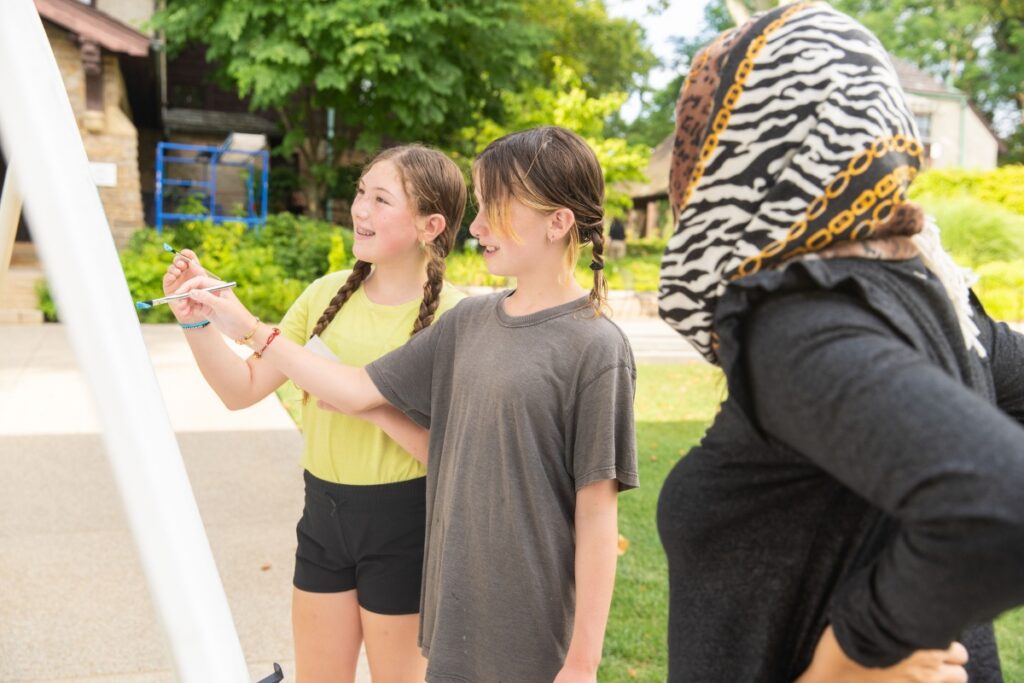 Two young girls with braided hair paint on an outdoor easel while an instructor looks on during a Summer Explorer Series artmaking program at The Frick Pittsburgh.
