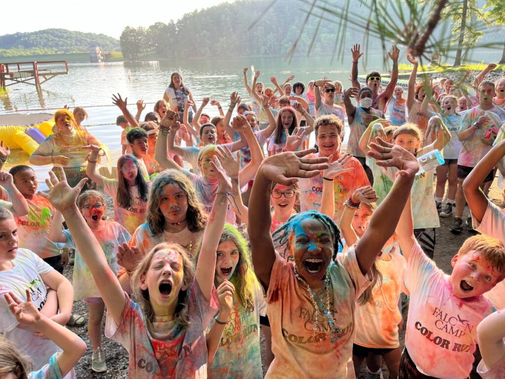 A large, vibrant group of boys and girls at Falcon Camp near Pittsburgh, covered in colorful paint powders from a recent color run, cheer and raise their hands in front of the scenic lakefront, celebrating the traditional summer camp experience.
