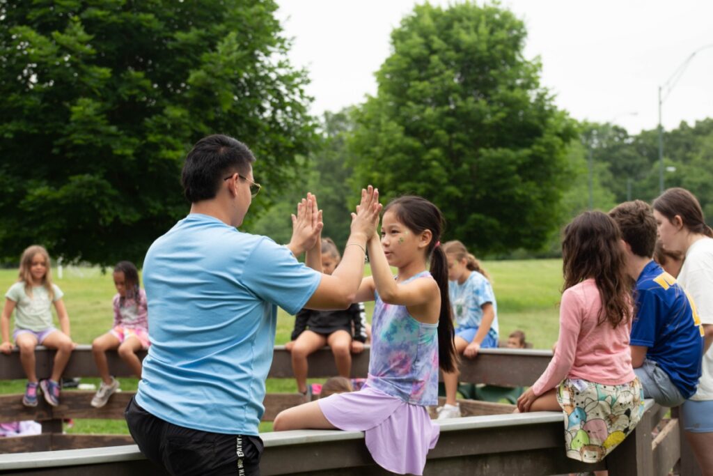 A young girl and a camp counselor high-five while sitting with a group of children at YMCA Camp Kon-O-Kwee Spencer.