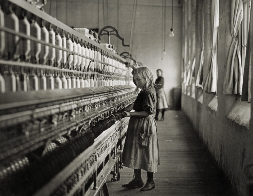 Historical black and white photograph by Lewis Hine of a young girl working at a spinning machine in a textile mill.