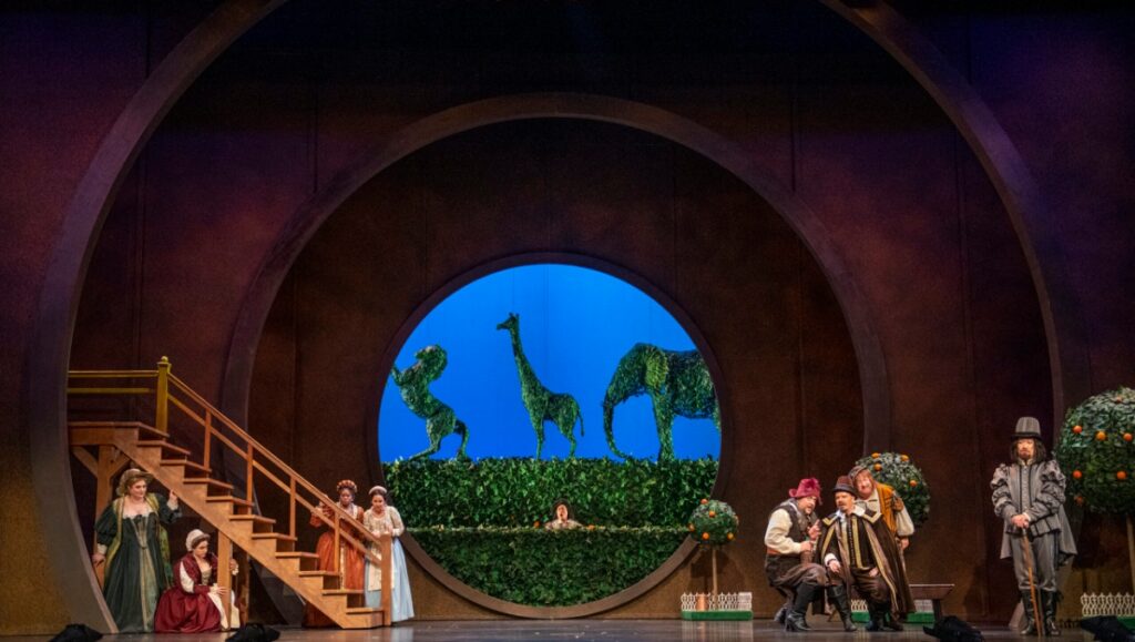 Stage production of Verdi’s Falstaff featuring opera singers in period costumes against a backdrop of blue topiary animals.