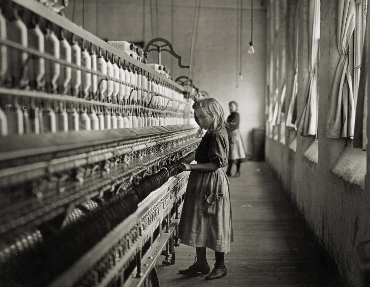Historical black and white photograph by Lewis Hine of a young girl working at a spinning machine in a textile mill.