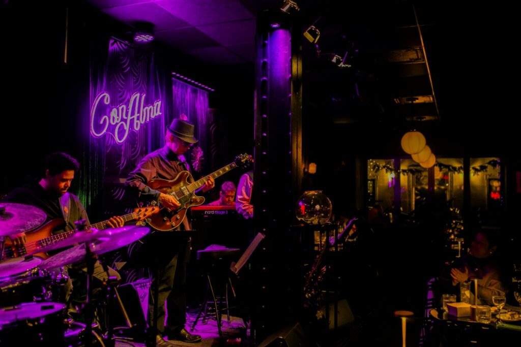 A live jazz performance at Con Alma in Downtown Pittsburgh, featuring a bassist and guitarist performing on stage under a glowing neon sign in a warm, intimate lounge setting.