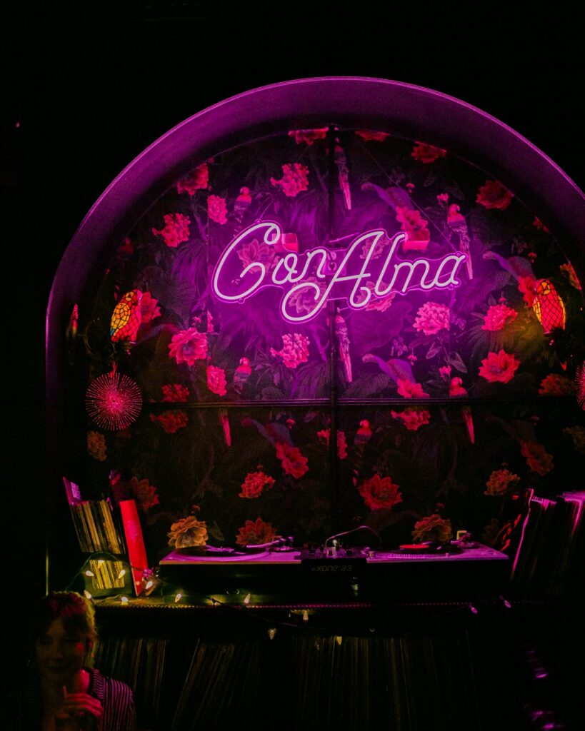 The pink neon light sign at Con Alma against a floral wall and DJ booth. 