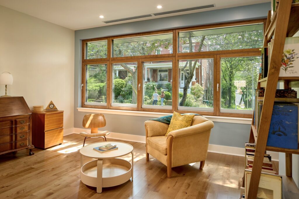 A yellow chair sits on the wood floor of a home living room by a wall of windows with wood frames.