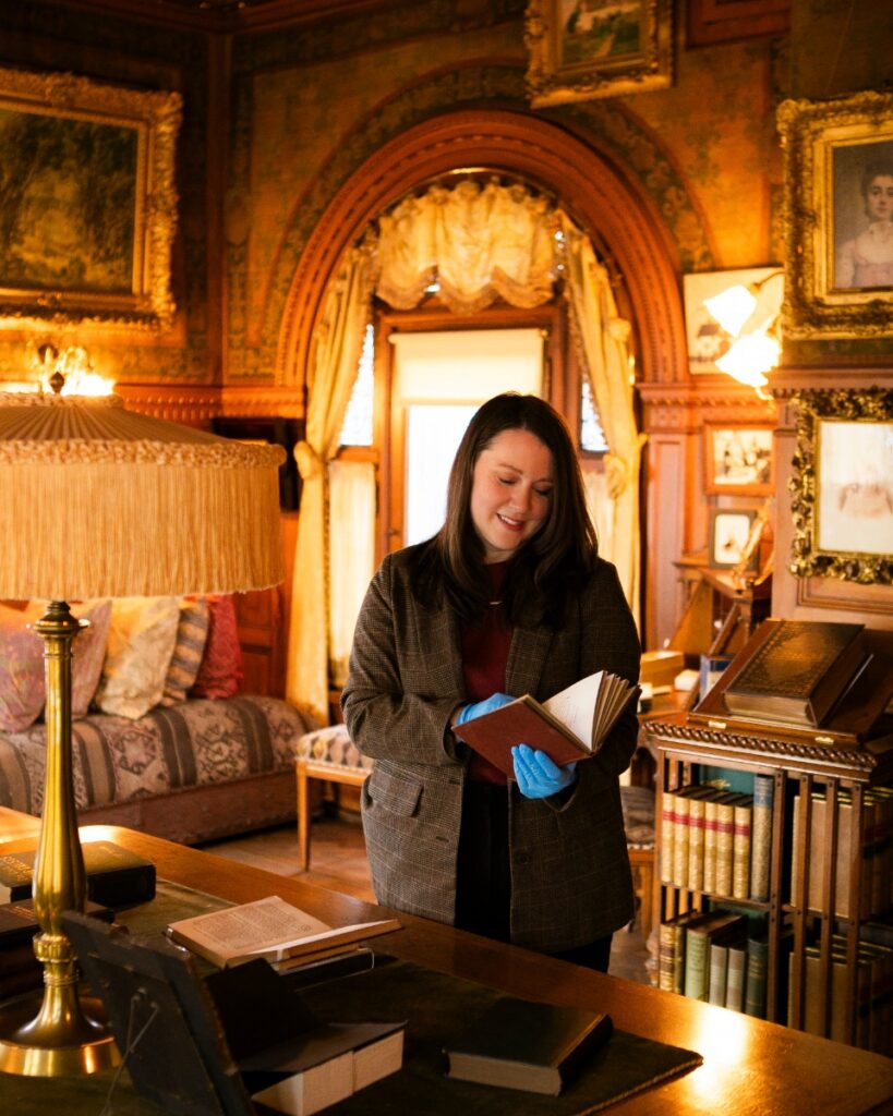Chief curator Dawn Reid Brean of The Frick Pittsburgh, wearing blue archival gloves, gently handles a rare book in the ornate, historic Clayton mansion library, surrounded by the Frick family's Gilded Age collection.