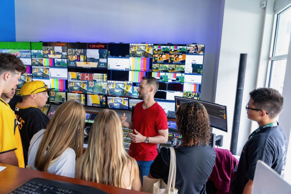 A man introduces various TV screens to a room of high schoolers. 