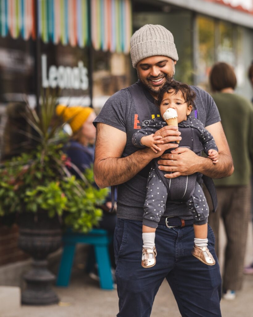A man holds a baby in front of Leona's ice cream as the baby licks from a vanilla ice cream cone. 