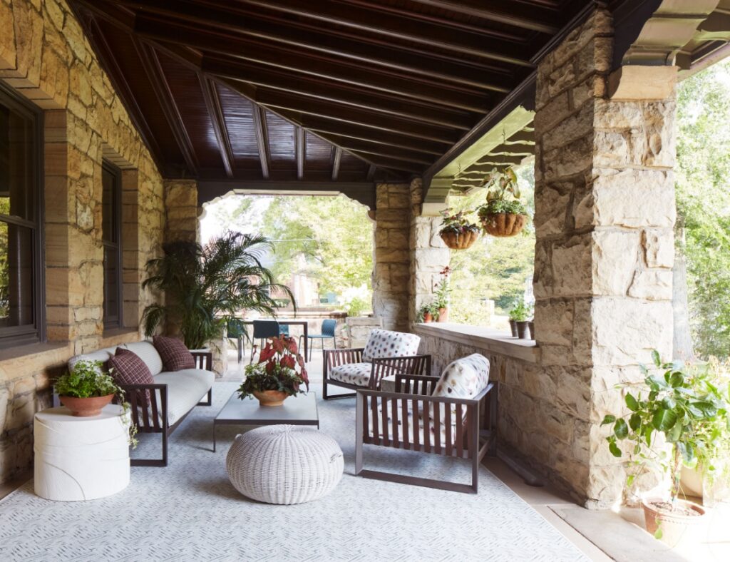 The spacious stone front porch of a historic Sewickley home features modern outdoor furniture, a dark wood-beamed ceiling, and lush potted plants, creating a welcoming and stylish outdoor living space.