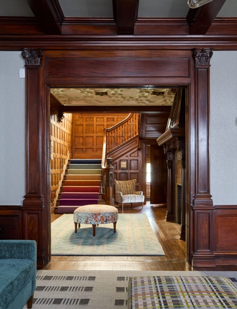 The grand foyer of a historic Sewickley home features a large wooden archway looking into an entry hall with a rainbow-colored staircase runner, an ornate plaster ceiling, and a floral-patterned ottoman.