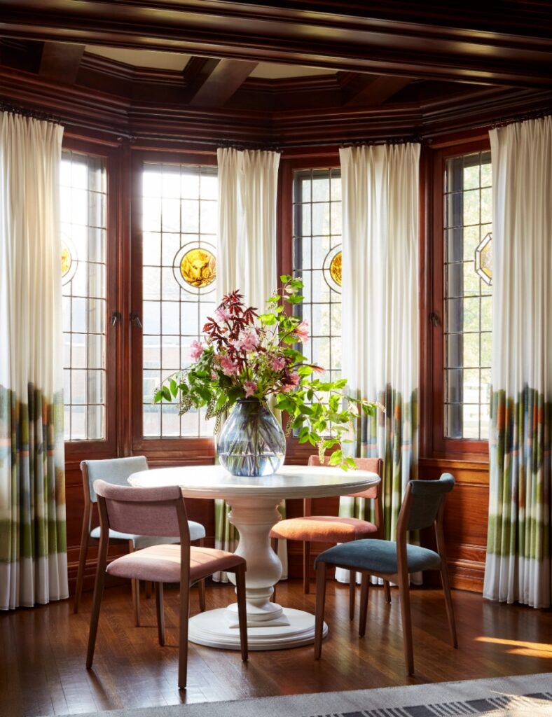 A bright, sunlit breakfast nook in a historic Sewickley home features a white pedestal table with colorful pastel chairs, set against a backdrop of ornate wood-paneled bay windows with stained glass details and landscape-patterned curtains.