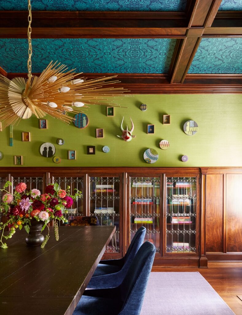 An eclectic dining room in a historic Sewickley home featuring a teal-patterned coffered ceiling, lime green textured walls adorned with small framed art pieces, and a striking gold starburst chandelier over a dark wood table with fresh flowers.