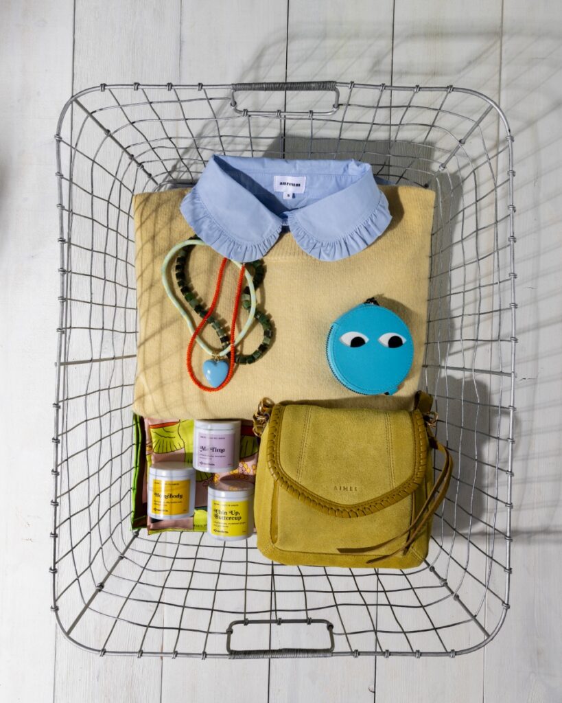 Curated artisan gifts and fashion in a metal wire basket, including a light blue sweater with a ruffled Peter Pan collar, beaded necklaces with a heart pendant, a blue round pouch with cartoon eyes, and three colorful candles.