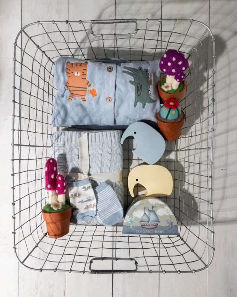 A curated wire shopping basket from Precious + Posh in Oakmont, filled with high-quality children’s items including an embroidered light blue top, a cable-knit blanket, infant socks, and whimsical felted potted plants.