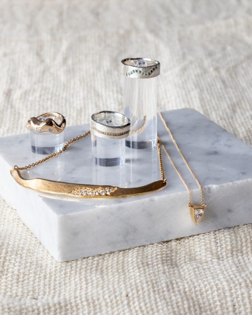 A variety of handcrafted fine jewelry from Suma Jewelry Co. in Oakmont, featuring sculptural gold and silver rings with blue and clear gemstones alongside a gold bar necklace and a triangle diamond pendant on a marble display.