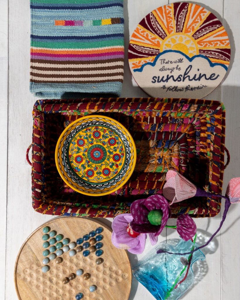 White plank floor featuring unique artisan gifts and global home decor, including a handcrafted mango wood Chinese checkers set with glass marbles, a "sunshine" embroidery hoop, and a patterned ceramic bowl.