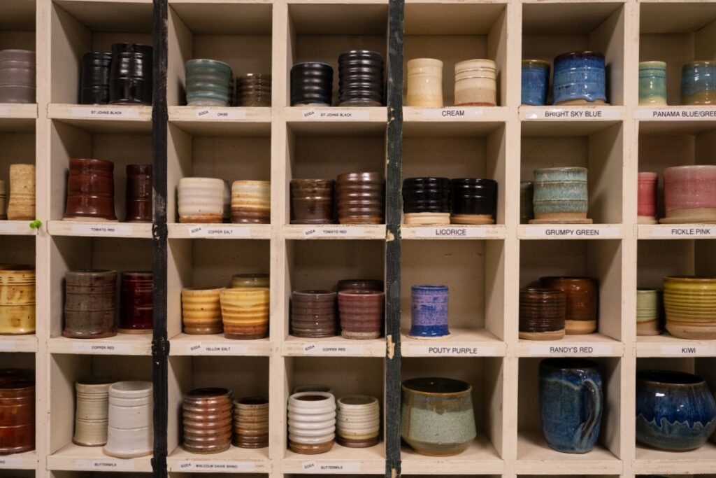 Shelves filled with colorful ceramic mugs and bowls, each labeled with names like "Cream" and "Pickle Pink," creating a vibrant, organized display.