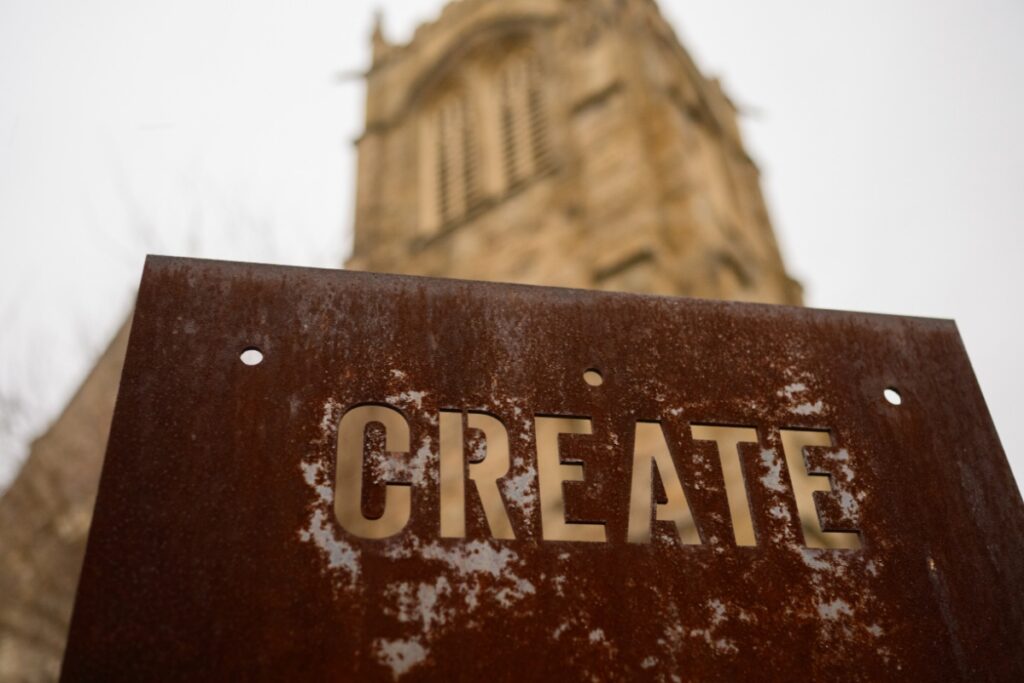 A rusted metal sign with the word "CREATE" cut out in stenciled lettering, with the blurred Gothic stone tower of the Union Project church visible in the background.