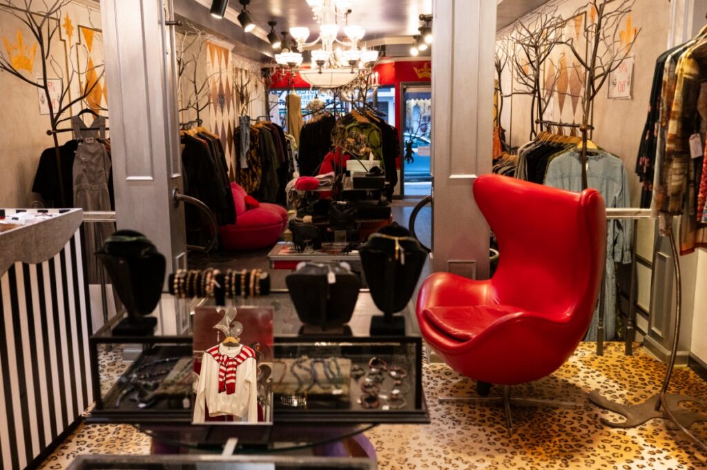 Chic boutique interior with a vibrant red chair, leopard print carpet, and racks of stylish clothes. A glass table displays jewelry and a sweater.