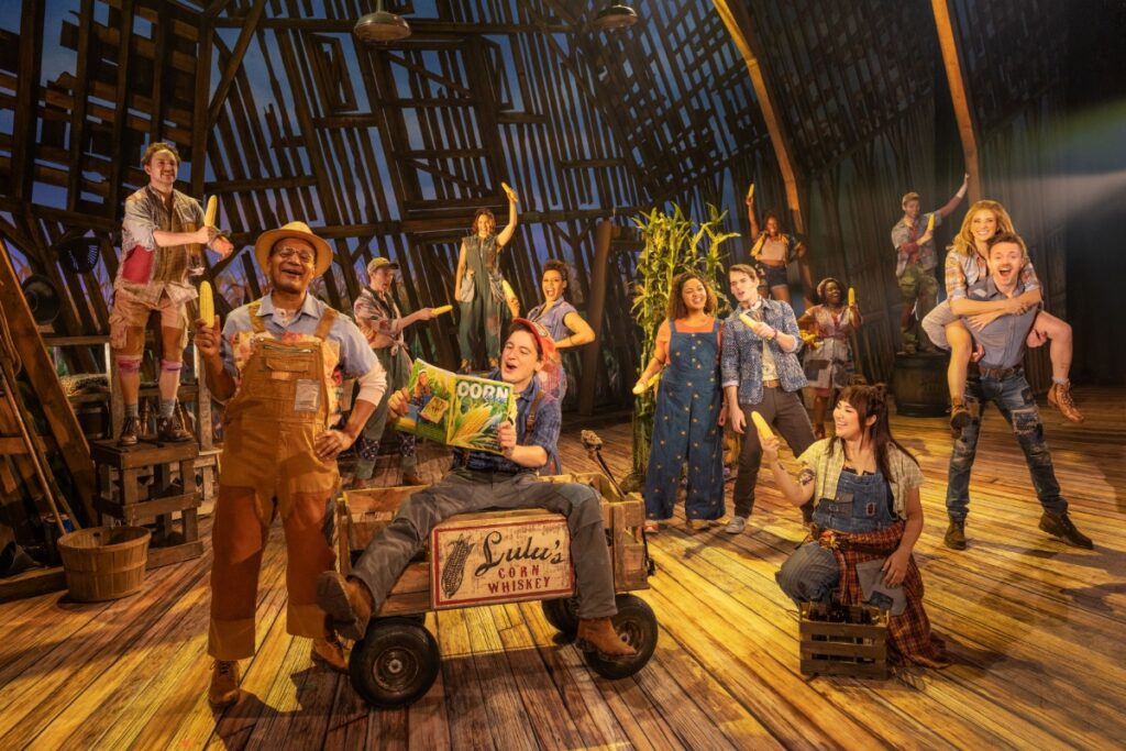 The cast of the Tony-winning musical "Shucked" performing a high-energy dance number in a rustic barn setting, featuring actors in colorful country-inspired costumes during the show's Pittsburgh debut.