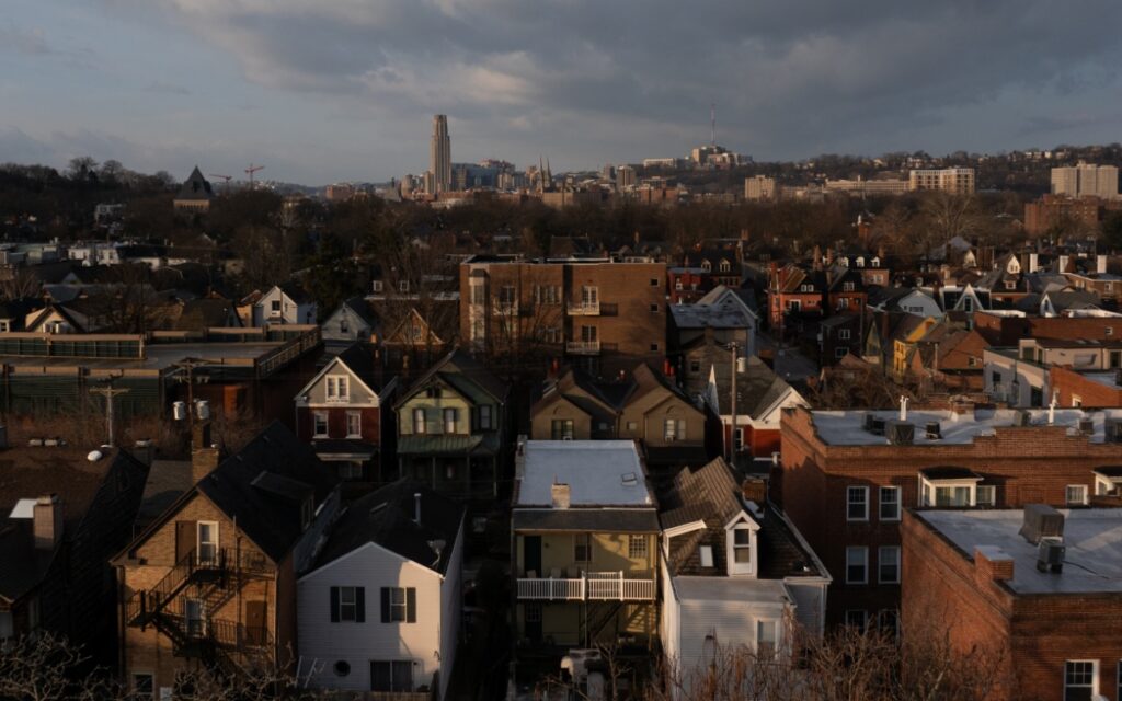 A Pittsburgh neighborhood at sunset, showing rows of historic residential houses and commercial buildings with the Cathedral of Learning and the city skyline visible on the distant horizon.