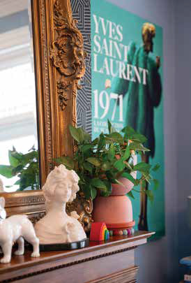 Detail of a historic wooden fireplace mantel decorated with a vintage white bust, green houseplant, and a large Yves Saint Laurent fashion poster.