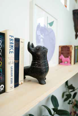 A minimalist light wood floating bookshelf styled with books, a black dog figurine, and a modern purple apple art print.