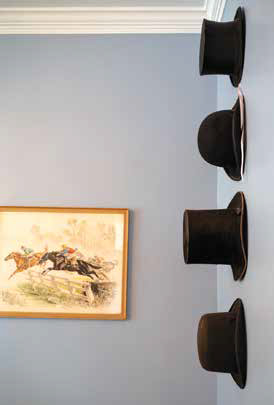 Antique black beaver top hats and wool bowler hats displayed vertically on a light blue wall next to a framed vintage horse racing print.