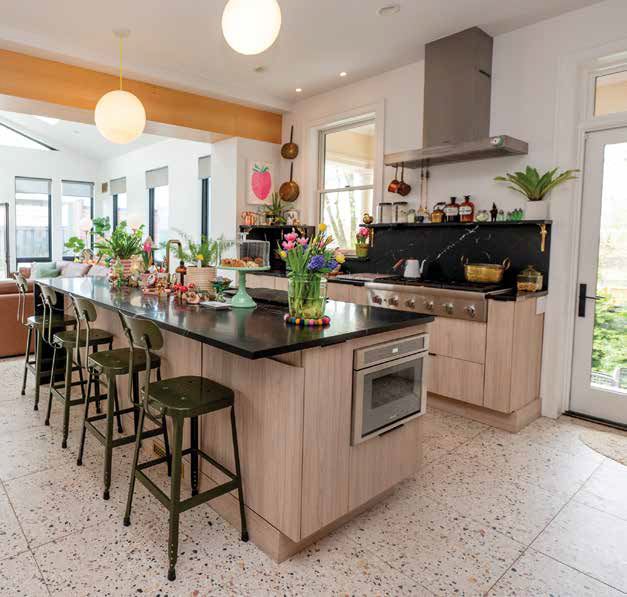 Modern kitchen with light wood cabinets, black marble countertops, a large island with green metal barstools, and terrazzo tile flooring.