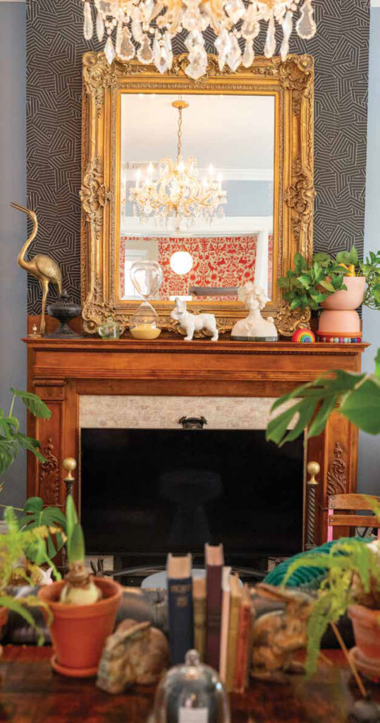 An ornate carved wood fireplace surround featuring a large gilded mirror reflecting a crystal chandelier and whimsical red and white folklore wallpaper.