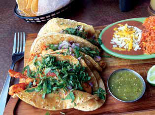 A platter of fresh street tacos topped with cilantro and onions on a wooden board, served with refried beans, rice, and salsa verde.
