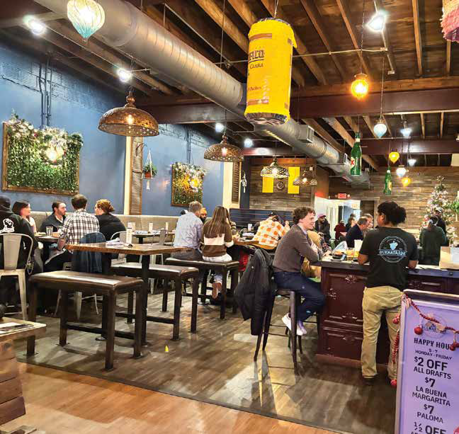 The lively, energetic interior dining room and bar area of Buenatarde restaurant with customers sitting under rustic hanging lights.