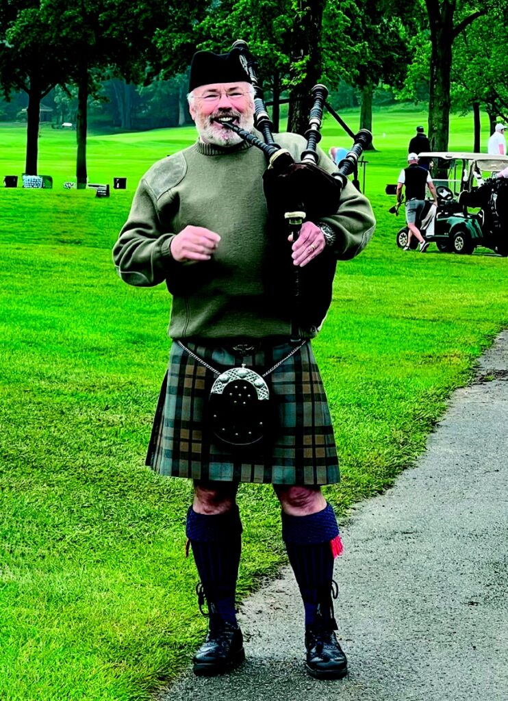 CMU Pipes and Drums member Walt McGhee dressed in a traditional kilt and Highland gear while playing the bagpipes on a golf course.