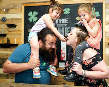 Owners Joe Bardakos and Madi Donaldson smiling in their restaurant with their two young daughters sitting on their shoulders.