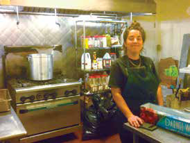 Sarah Coppolo wearing a dark apron in the commercial kitchen of Brothmonger next to a large stainless steel soup pot on a gas stove.