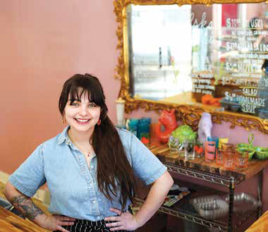 Owner Olive Visco smiling in her Polish comfort-food café, Polska Laska, in front of a vintage gold mirror displaying the daily menu.