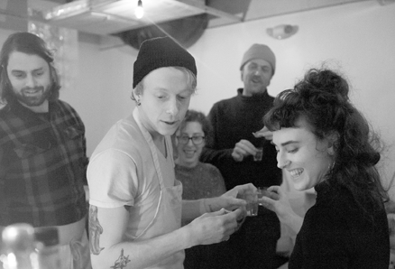 A candid black and white photo of a group of friends smiling and raising a small glass toast together at a pop-up dinner.