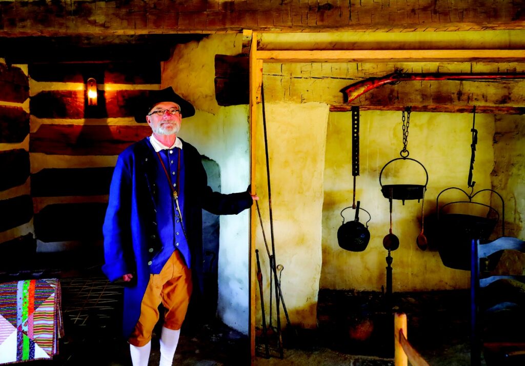A park docent dressed in traditional 18th-century attire standing inside the Neill Log House next to the original stone chimney.