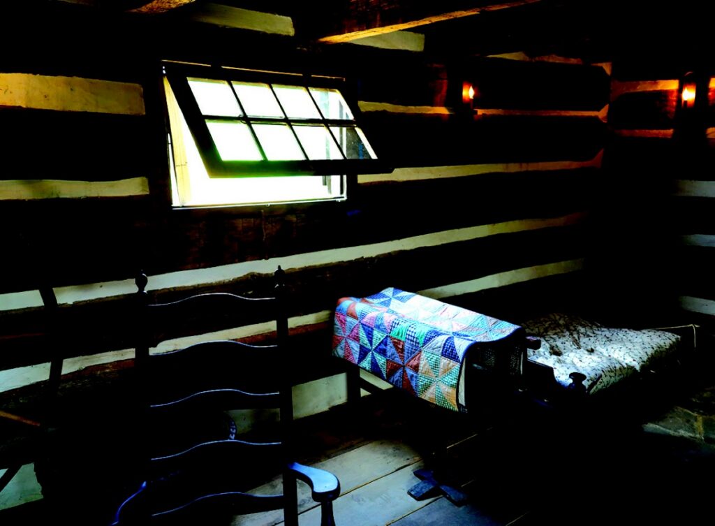 The interior loft of the Neill Log House showing a traditional string bed covered with a colorful patchwork quilt near a small glass window.